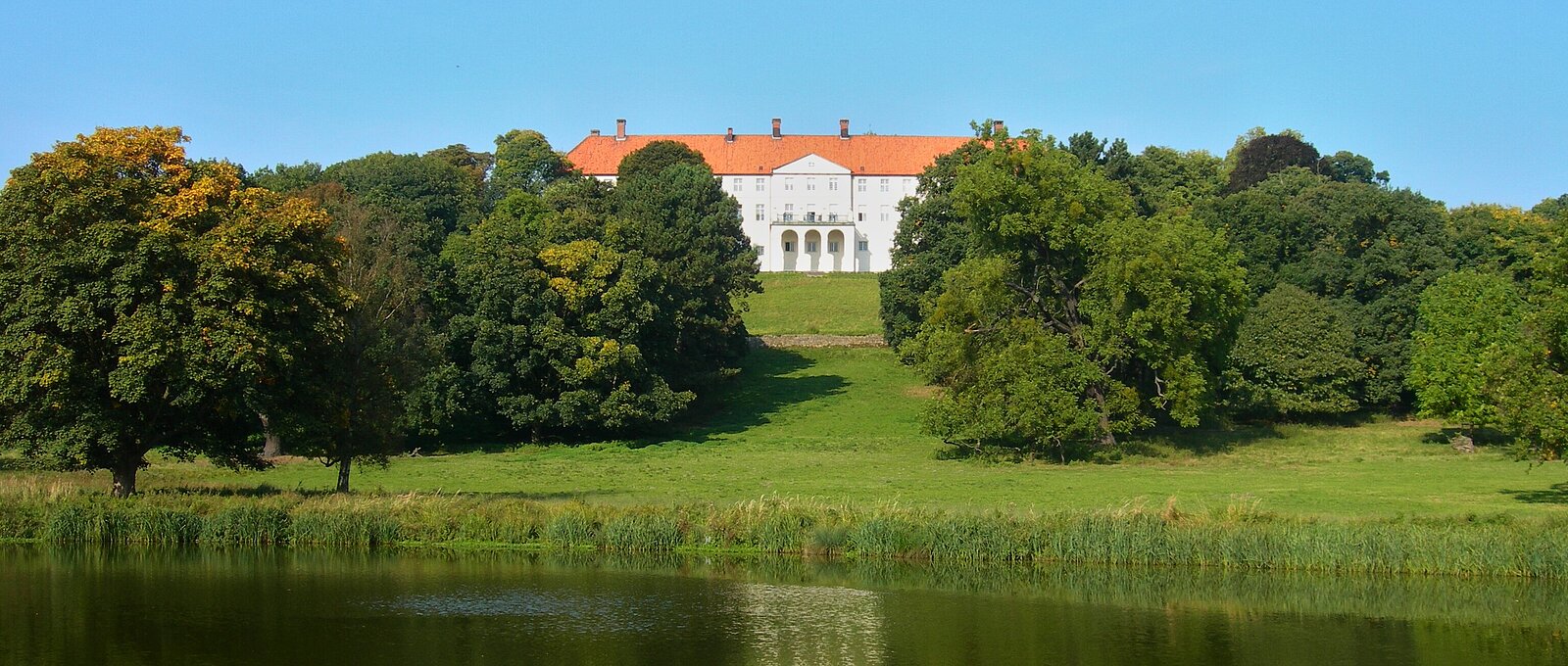 Weißes Schloss mit rotem Dach auf Anhöhe hinter See