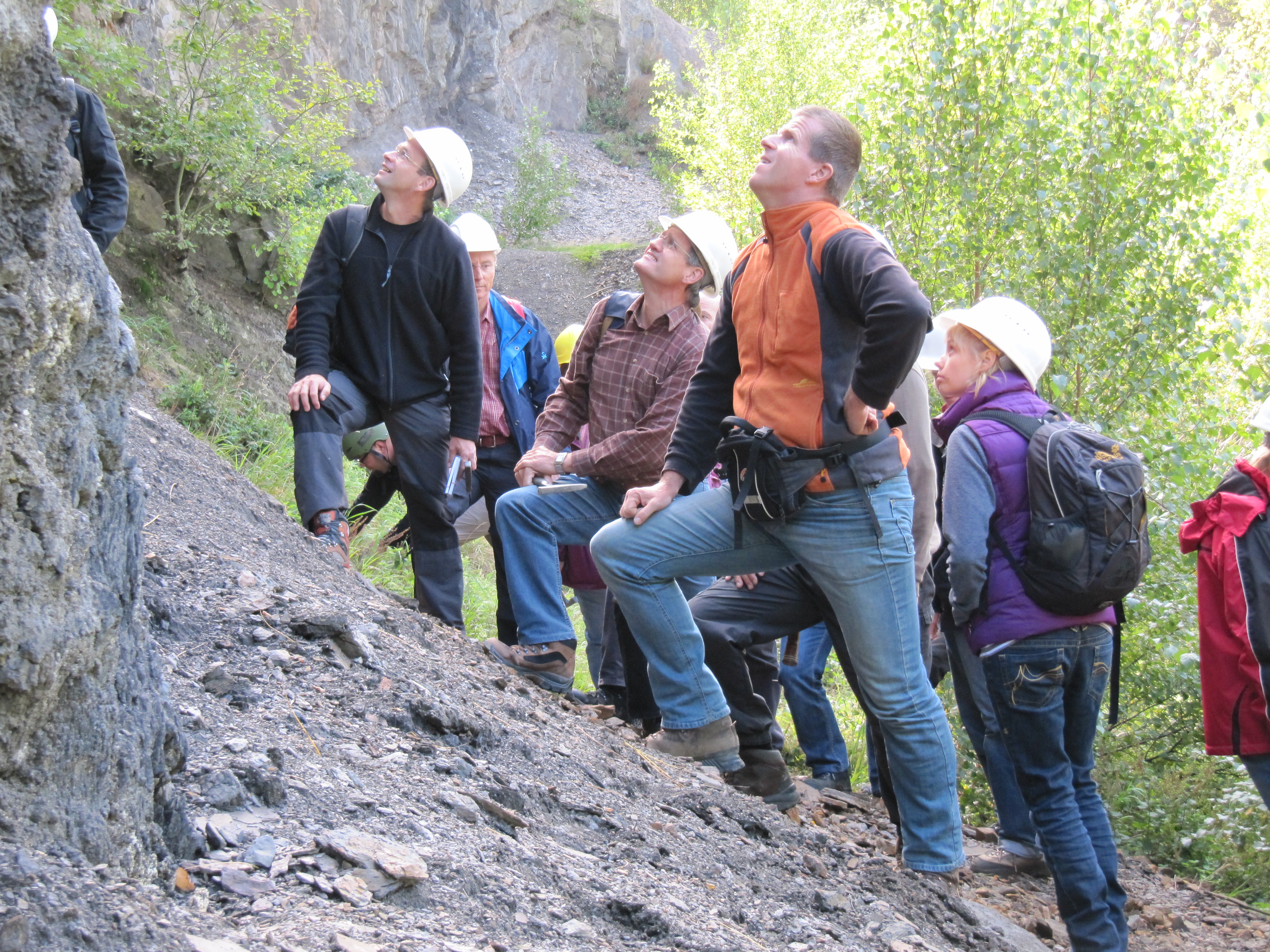 Erwachsenengruppe mit Helmen an Abhang vor Felswand.