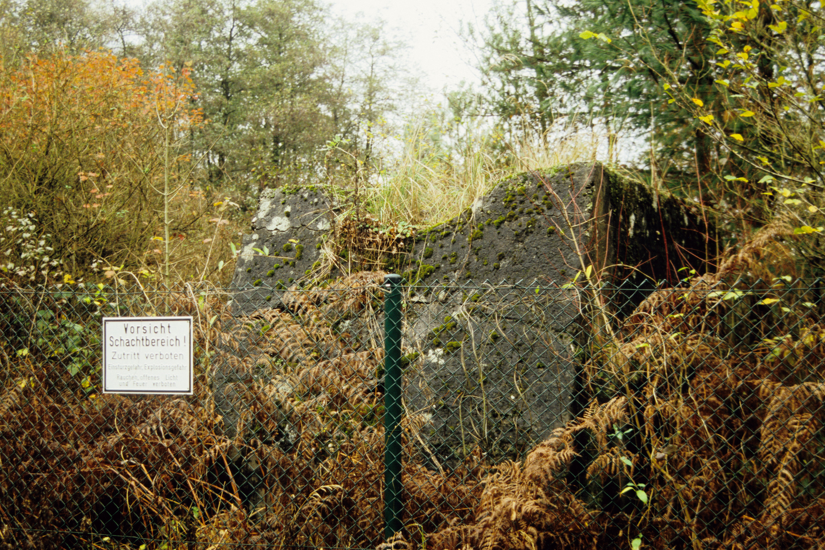Zaun mit Schild "Vorsicht Schachtbereich" und Gebäuderest